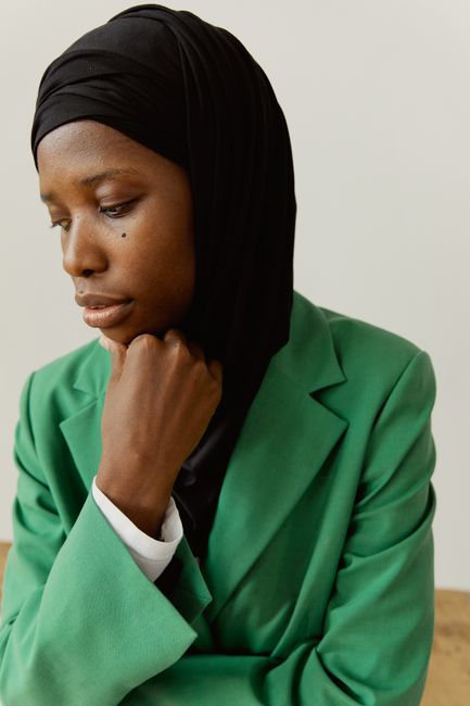 A pensive woman wearing a green blazer and black hijab, posing with a hand on her chin indoors.