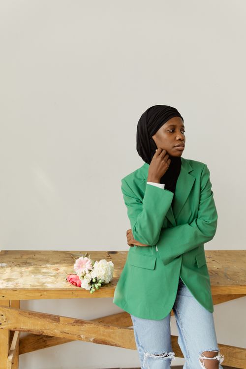 Free Woman in green blazer and hijab stands by a wooden table with flowers. Stock Photo