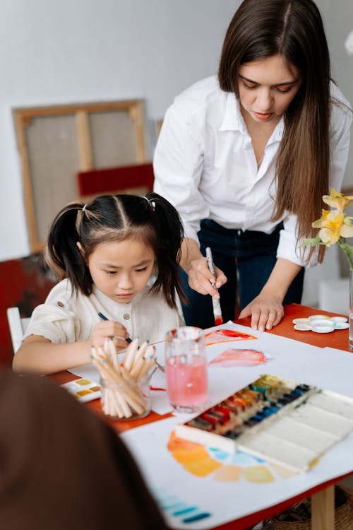 A Woman in White Long Sleeves Standing Near Her Student Painting on White Paper