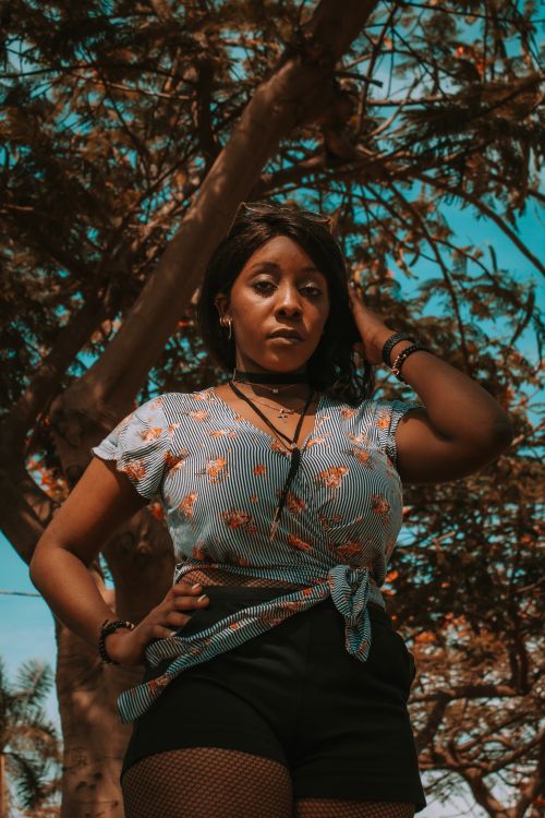 Free African American woman posing confidently under a tree wearing a floral blouse and black shorts. Stock Photo