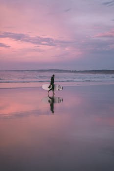 A surfer carrying a surfboard walks along the tranquil shoreline at sunset, reflecting in the water.