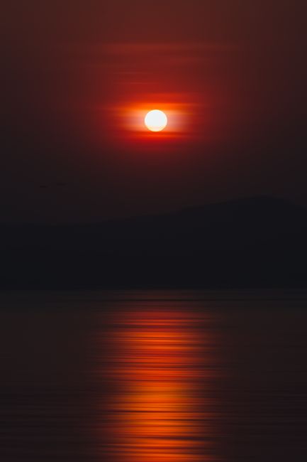 Vibrant sunset over water with striking reflections and silhouette of mountains.