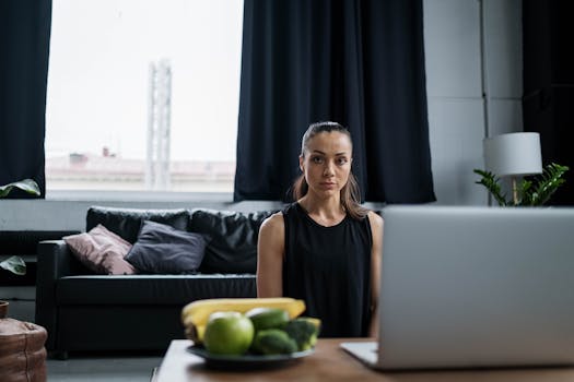 Woman in living room with laptop, sitting at table with fruits and vegetables nearby.