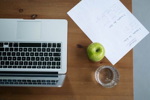 Free Top view of a workspace with a laptop, green apple, water glass, and paper notes. Stock Photo