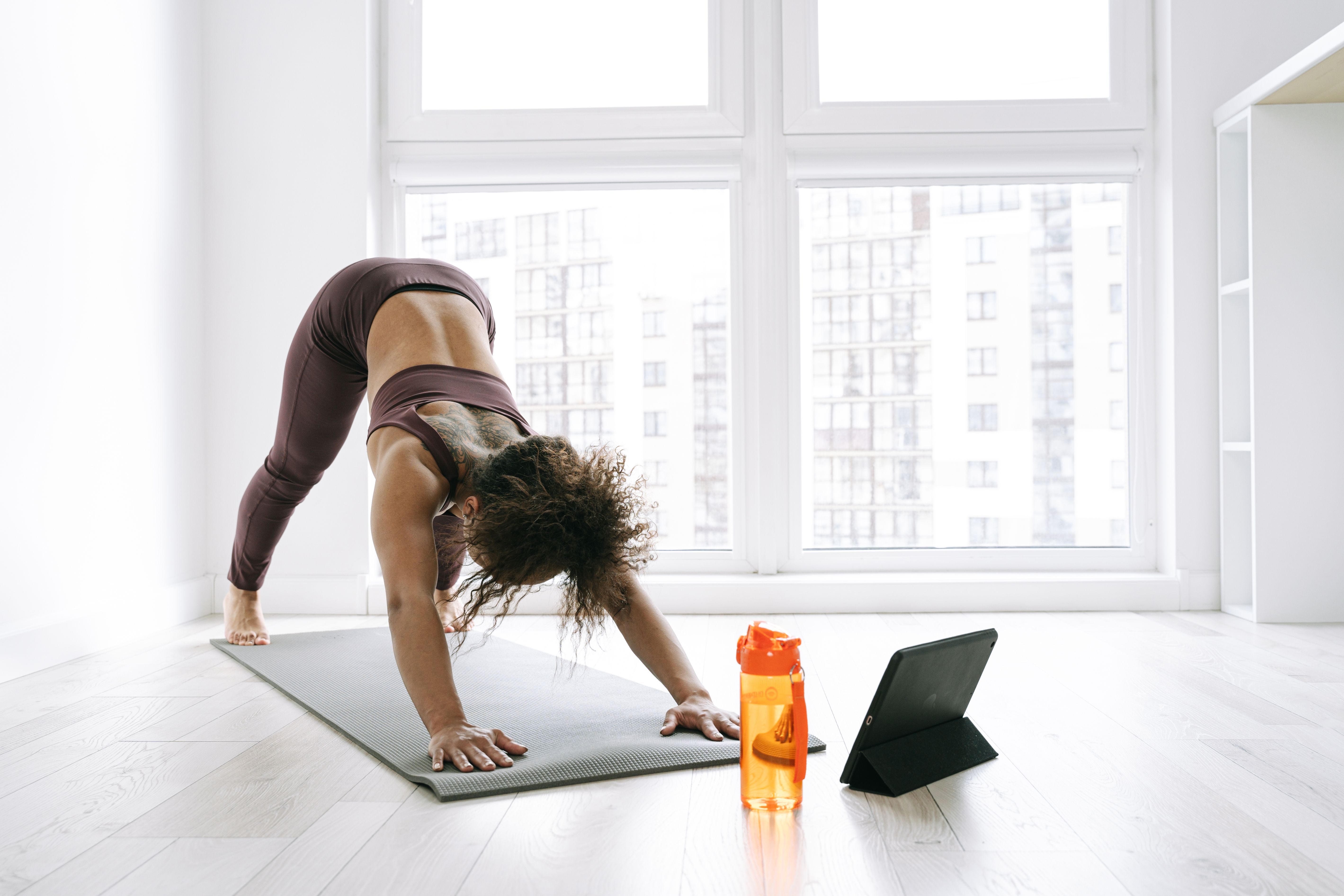 A woman doing yoga in a sunny room with a tablet and water bottle nearby, emphasizing a healthy lifestyle.