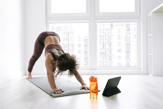 A woman doing yoga in a sunny room with a tablet and water bottle nearby, emphasizing a healthy lifestyle.