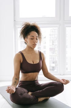 Adult woman meditating in a yoga pose on a mat indoors, promoting fitness and relaxation.
