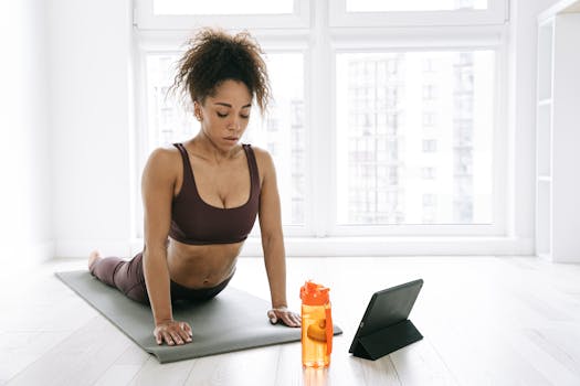 A woman in activewear performs yoga on a mat indoors, next to a tablet and water bottle.