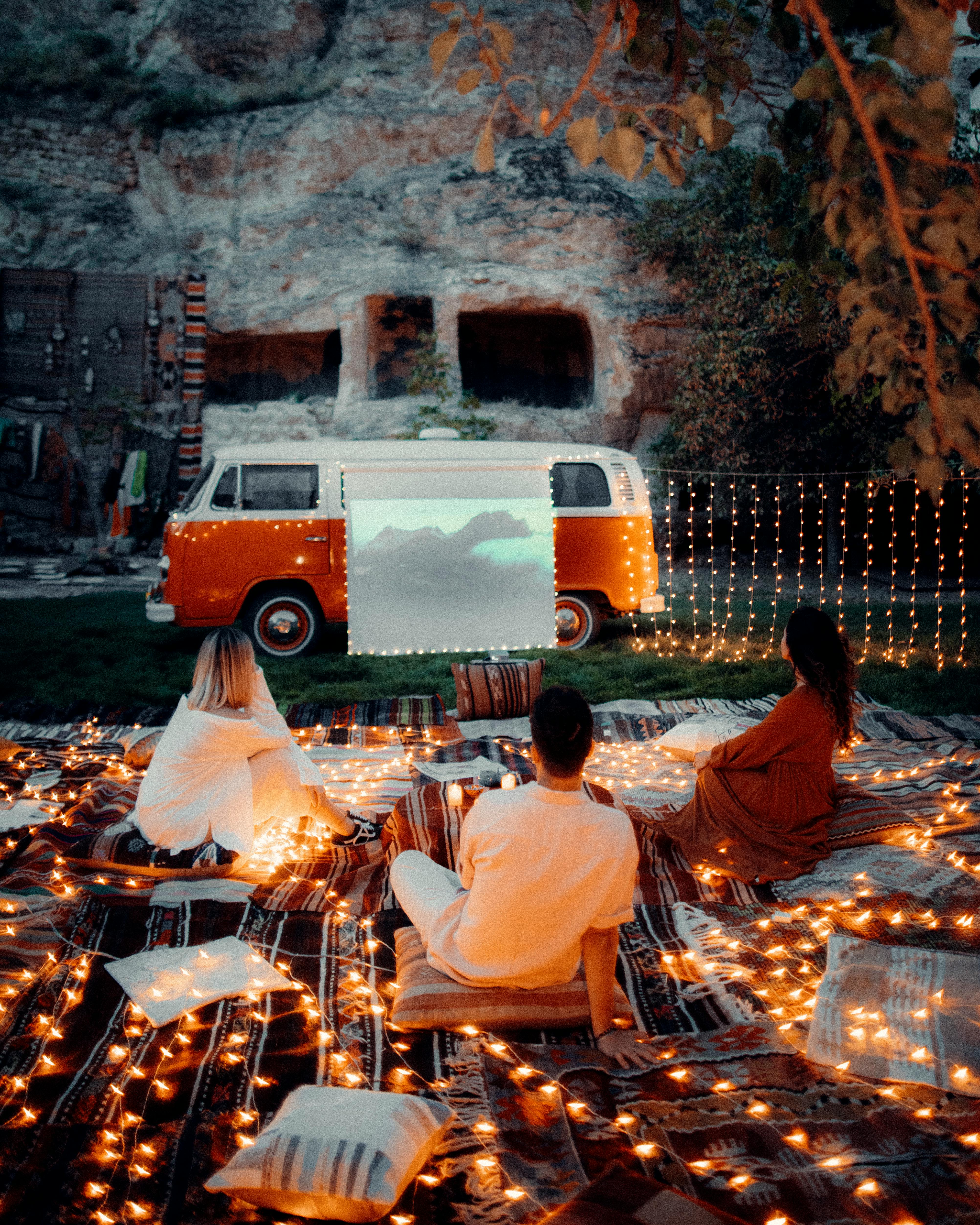 Back view of unrecognizable friends sitting on cushions near camping van and rocky hill while watching film in evening