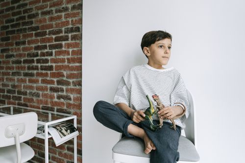 Free Child sitting on chair, holding dinosaur toys, in stylish indoor setting with brick wall. Stock Photo