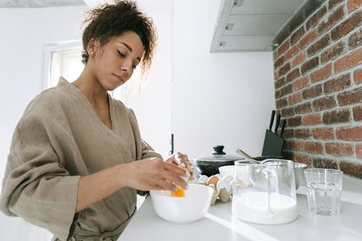 Stylish young woman cracking an egg into a bowl while cooking in a bright modern kitchen.