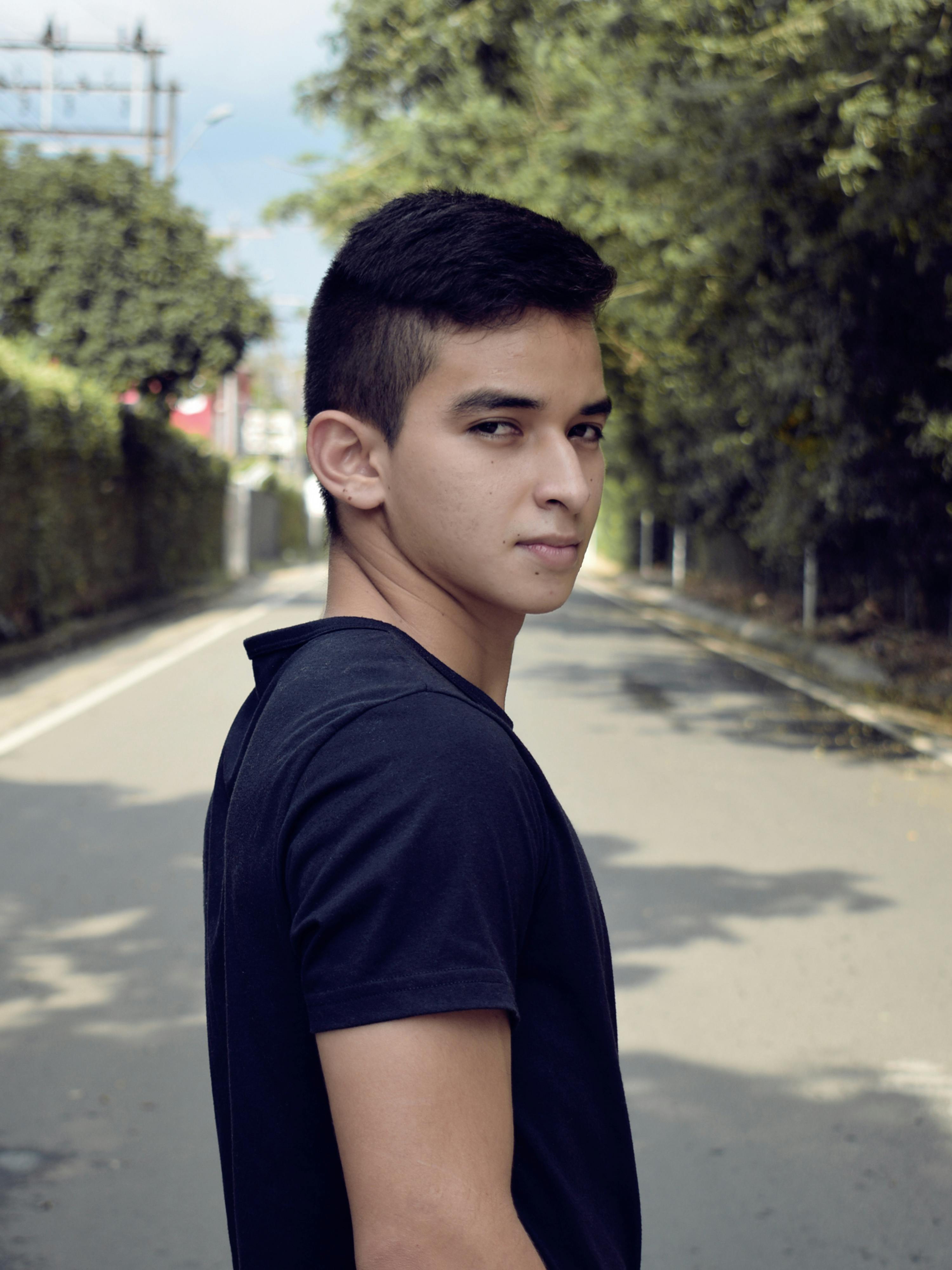 Young man wearing a black shirt posing confidently on a serene tree-lined street.