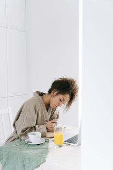 Woman in a cozy robe writes notes on a table with juice and laptop at home.