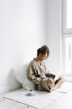 A woman sits on a rug in a bathrobe using a laptop, enjoying a cup of coffee.