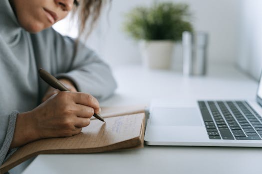 A woman writes in a notebook at a desk with a laptop, personifying remote work.