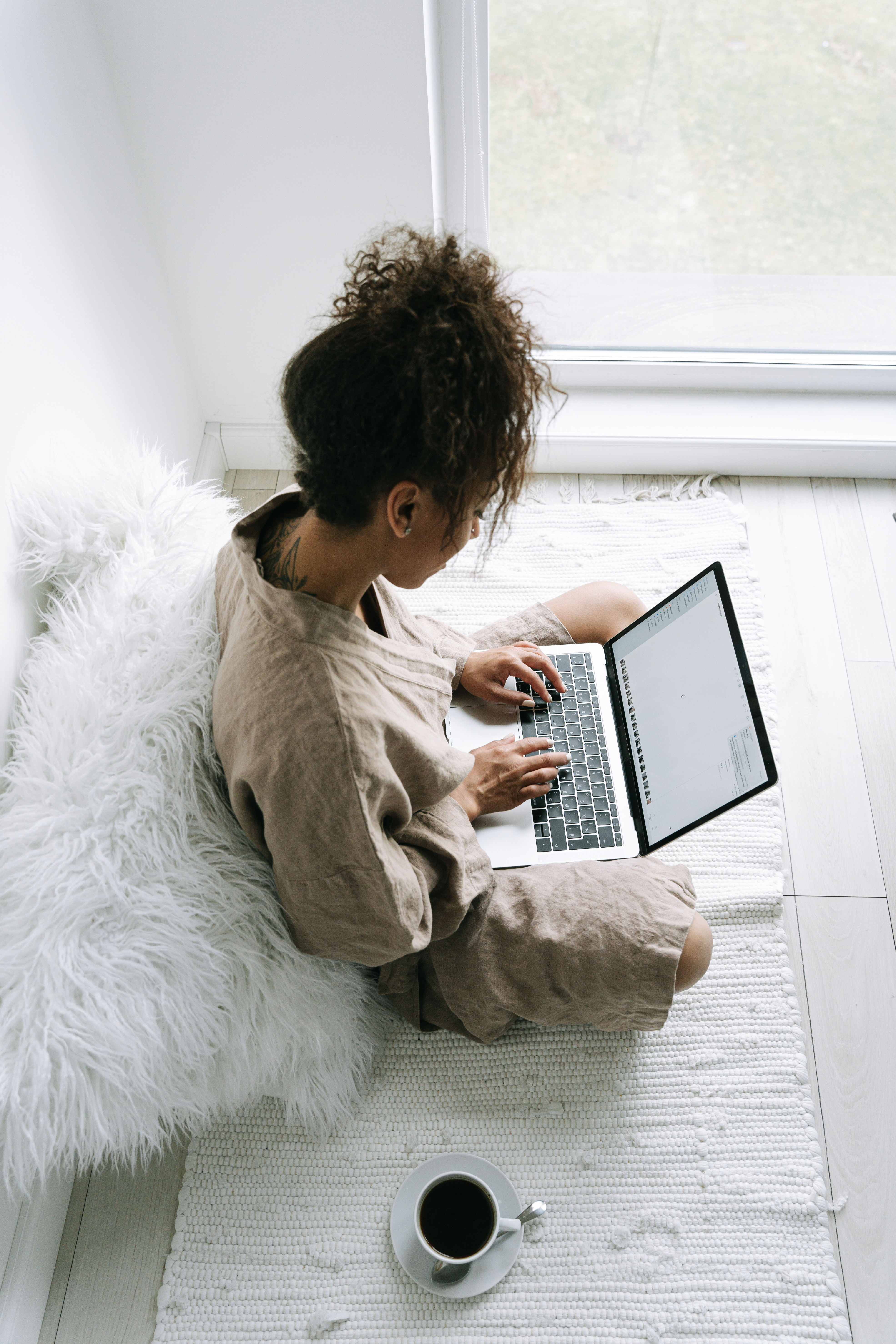 Woman in a cozy bathrobe using a laptop, sitting comfortably on a rug beside a window.