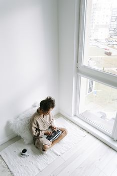 African American woman in robe working on laptop by window, conveying remote work lifestyle.