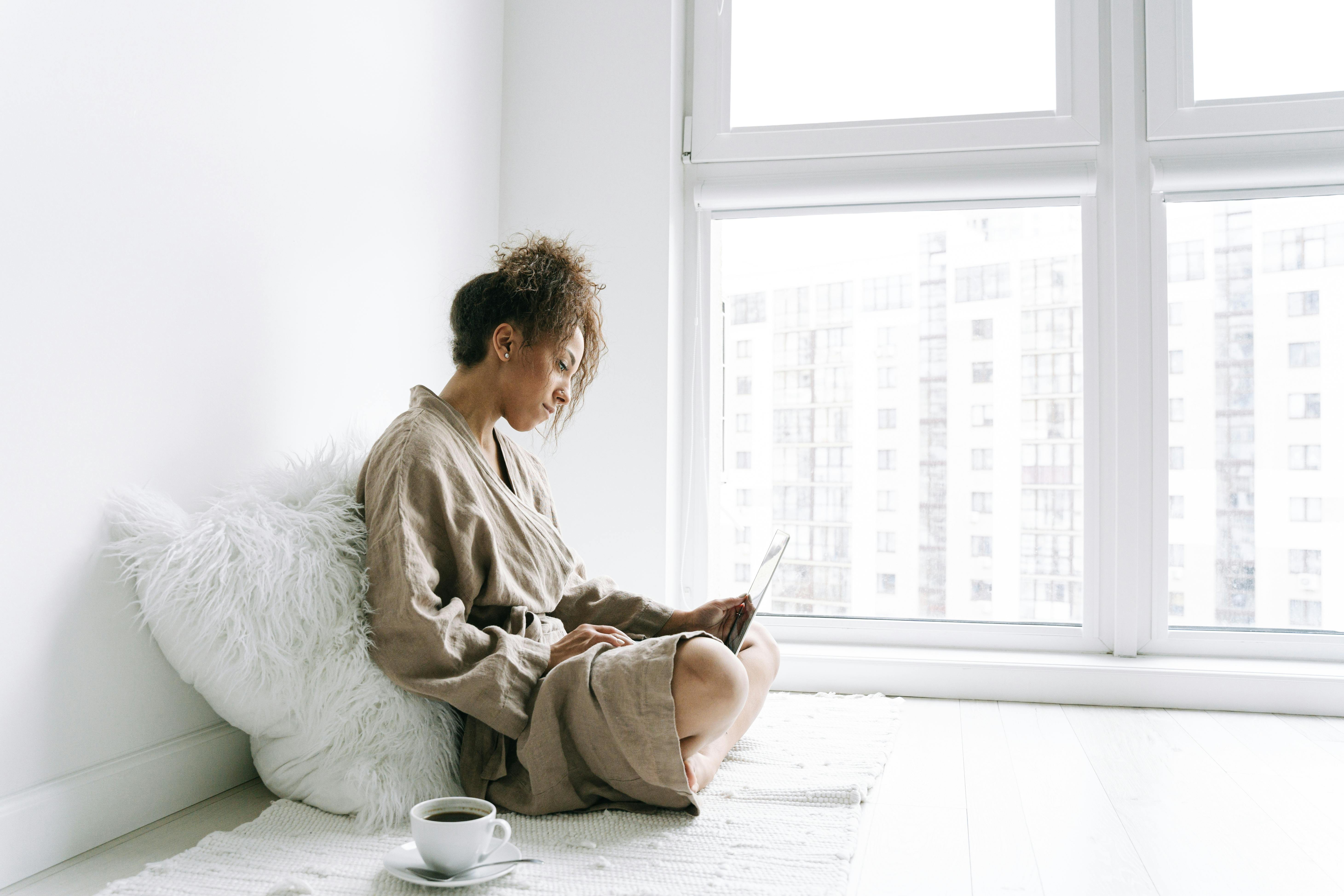 A woman in a robe sits by a window, using a laptop with a cup of coffee nearby