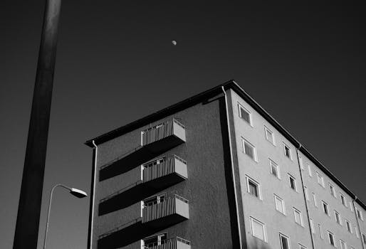 A dramatic black and white low-angle view of a city building under a clear sky with the moon visible.