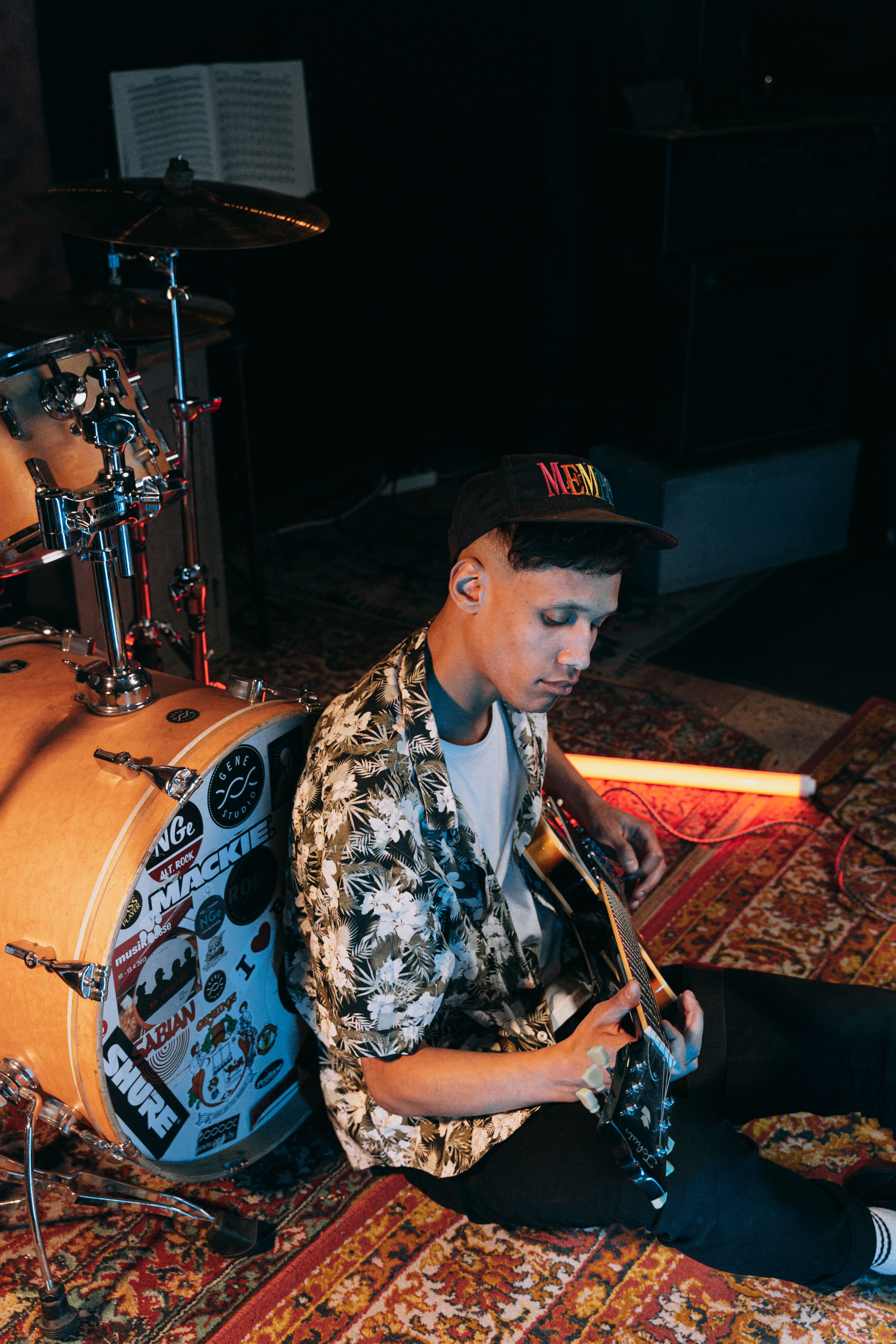 A young man sitting on a studio floor playing an electric guitar next to a drum set.