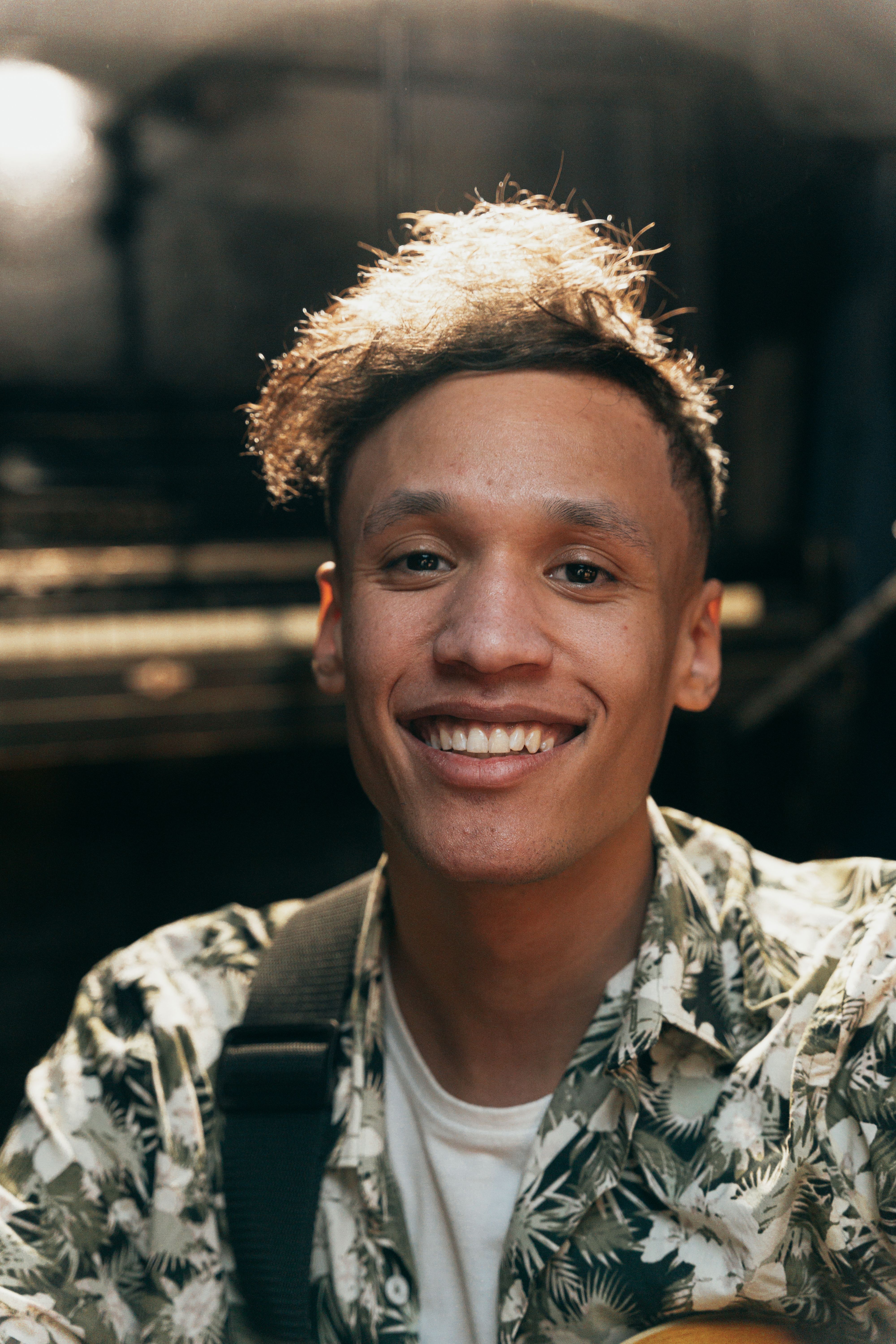 Portrait of a smiling man in a floral shirt in a warmly lit indoor setting.