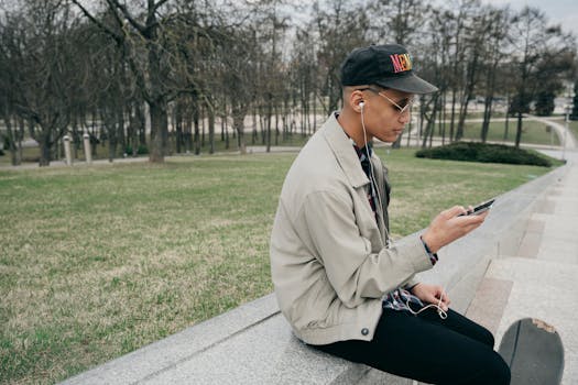 A stylish young man sitting on a bench, listening to music on a smartphone outdoors.