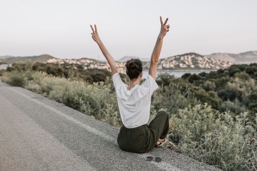 Woman on a roadside in Antalya raises peace sign, embracing summer with mountain views and tranquility.