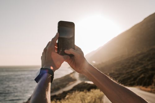 Free Person taking a photo of the sunset over the coastline with a smartphone. Stock Photo