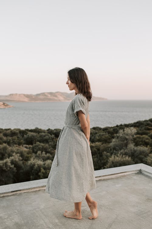 Woman in Gray Dress Standing on Gray Concrete Looking at the Sea