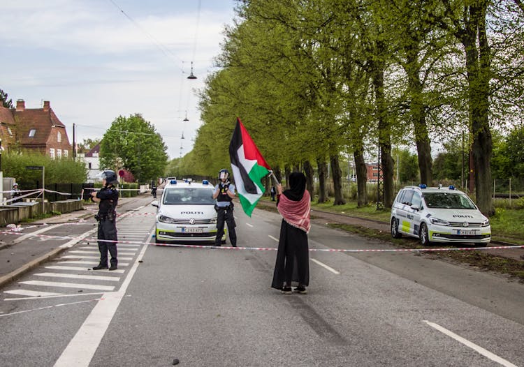 A Person In Black Dress And Hijab Waving Flag In Front Of A Police