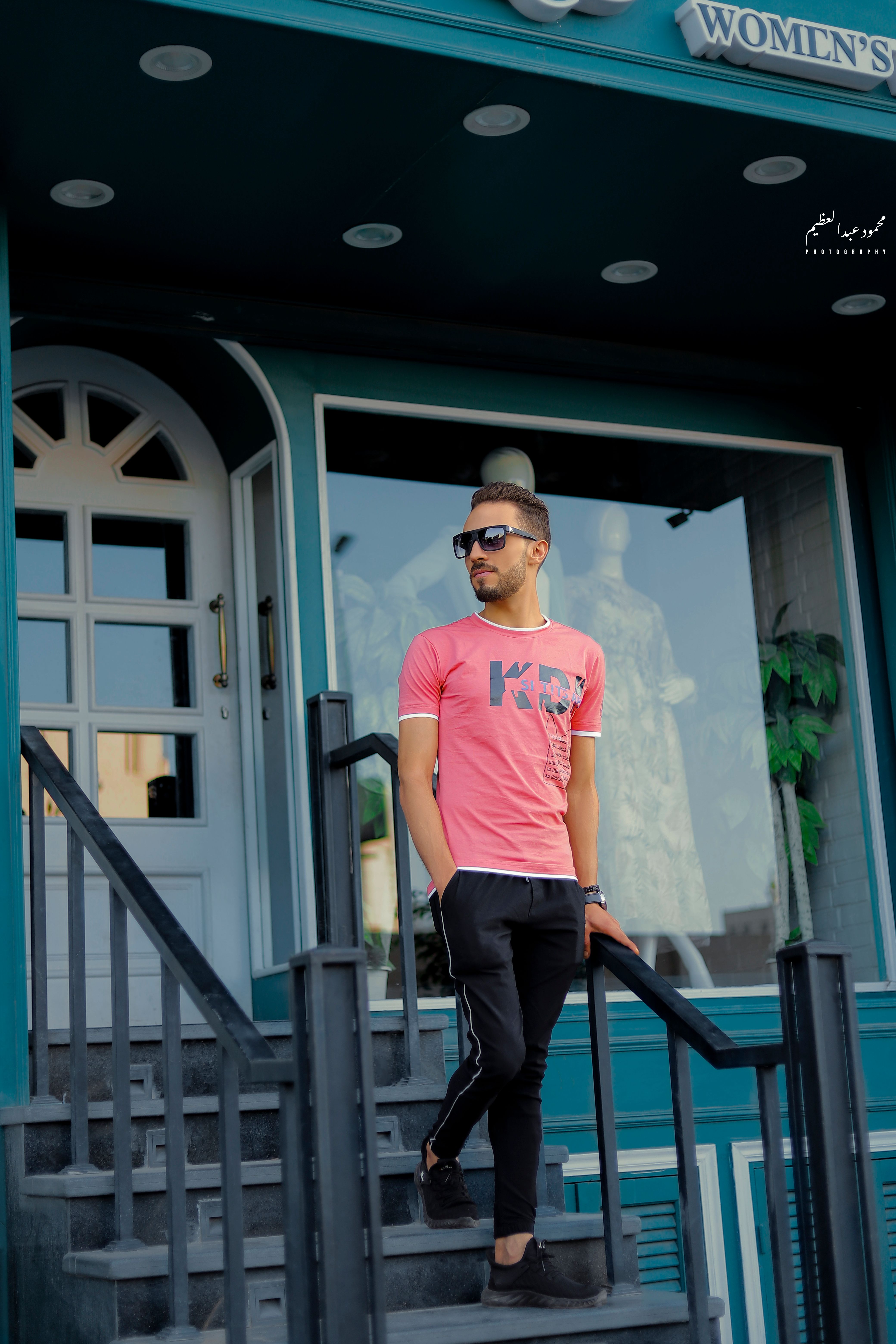 Fashionable man in sunglasses and pink shirt standing on urban staircase outside shop.