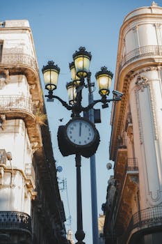 Elegant vintage clock and street lamps nestled between historic buildings under a bright sky.