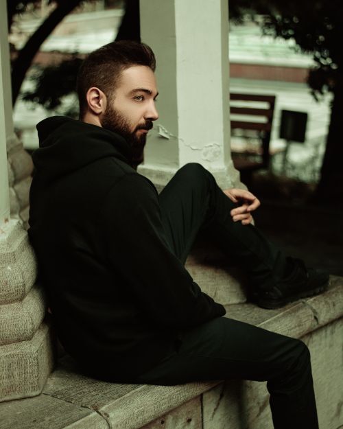 Free A man with a beard sitting on a stone bench outdoors, wearing a black hoodie. Stock Photo