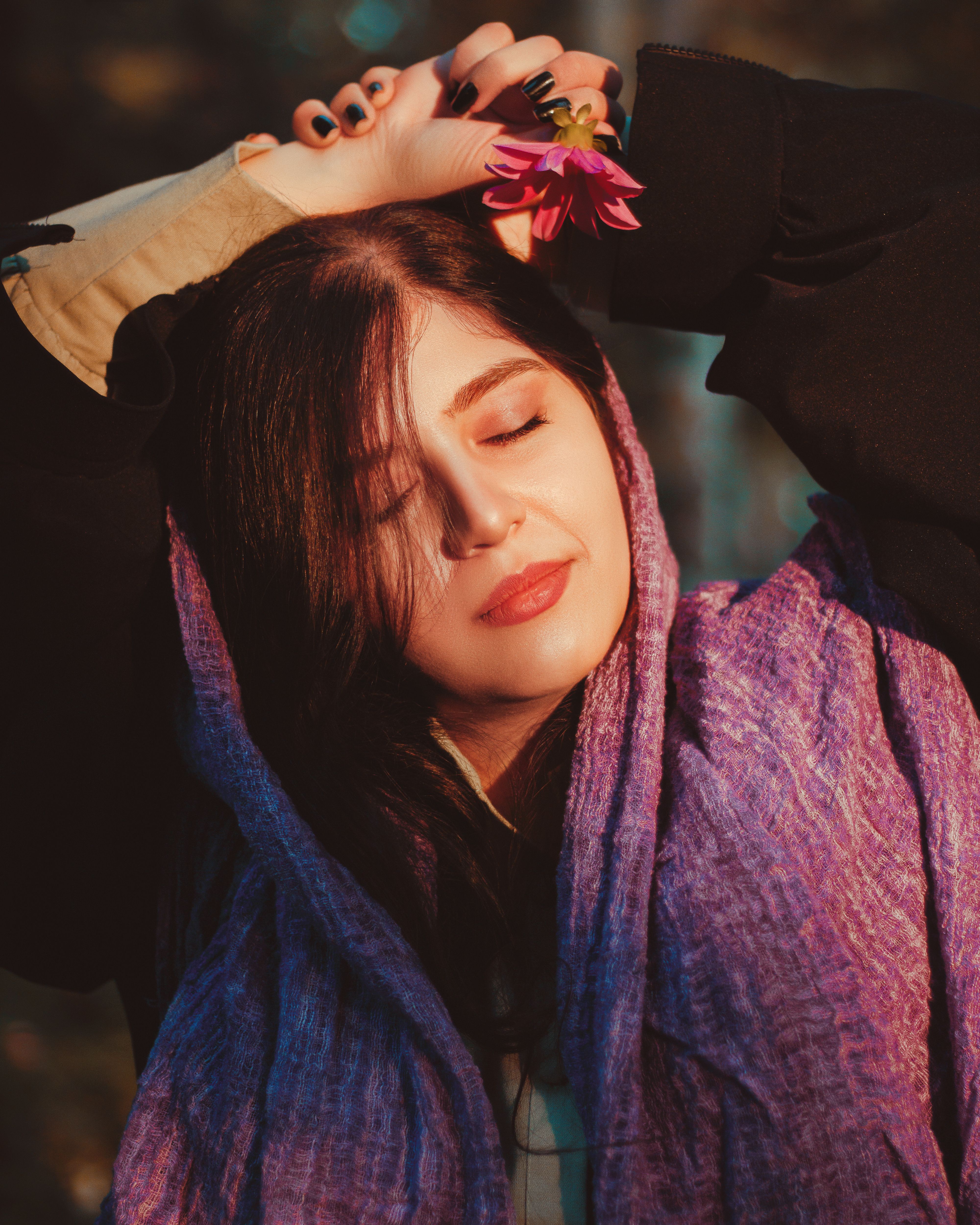 A serene woman with eyes closed holds a flower outdoors, basking in the warm sunlight.