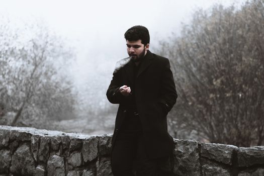 Portrait of a man smoking by a stone wall in a foggy outdoor setting.