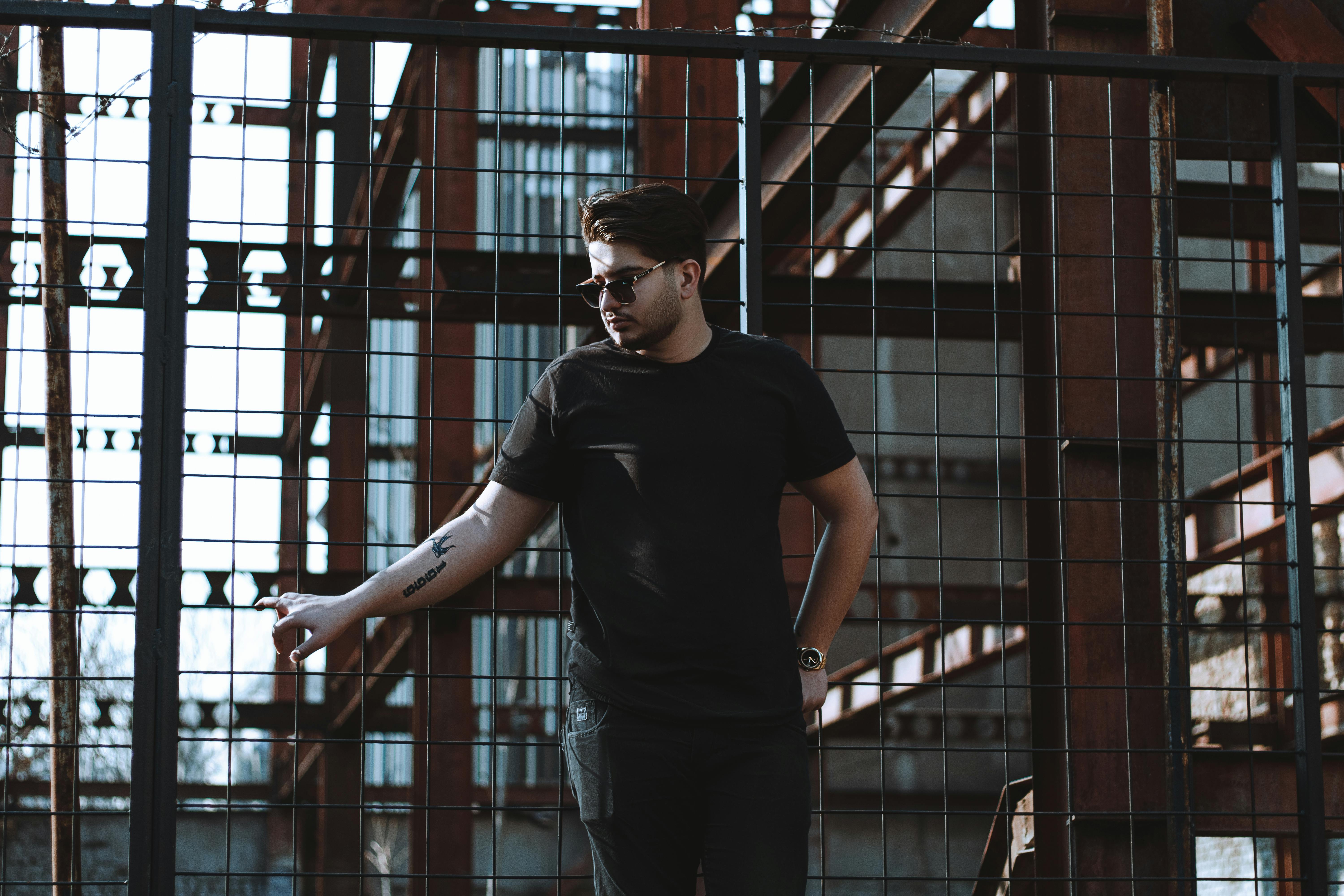 A young man in sunglasses and black t-shirt stands stylishly against a metal fence in an industrial setting.