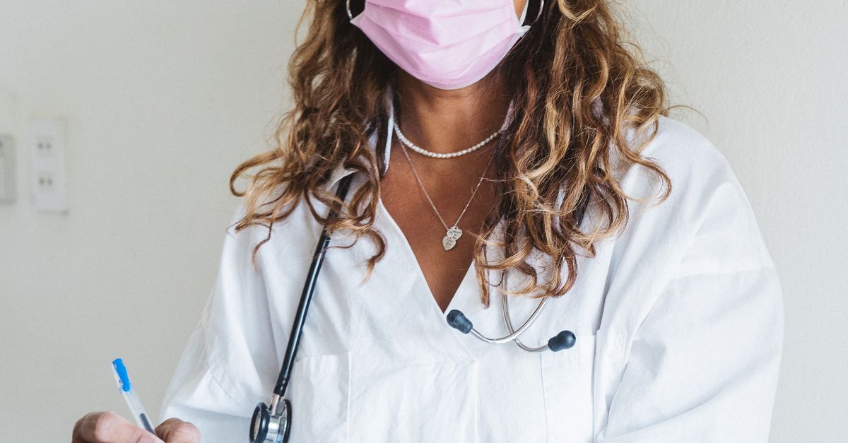 Photo by Anna Tarazevich A female doctor in a lab coat, writing in patient records, wearing a pink surgical mask, indoors.