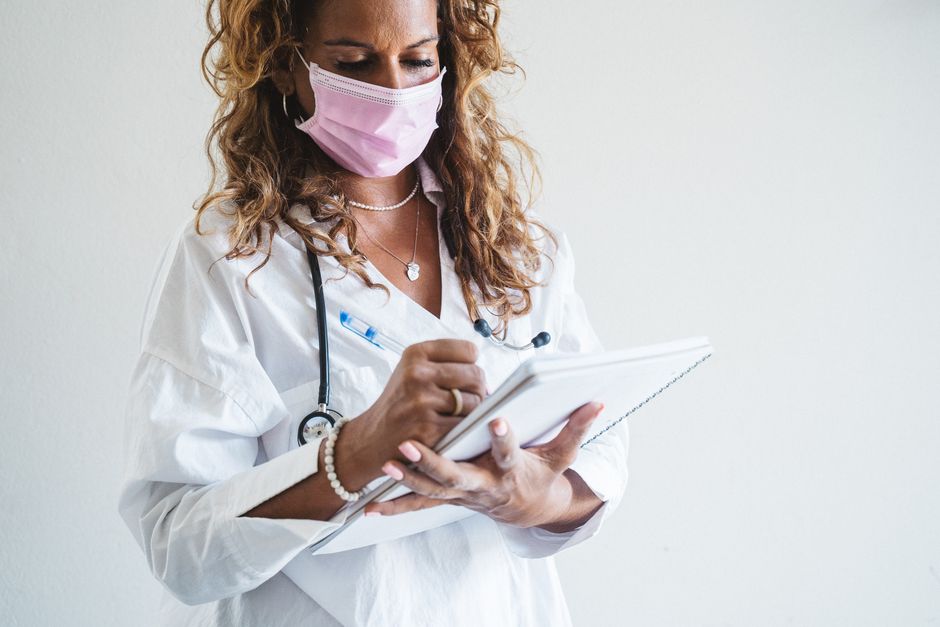 A female doctor in a mask writing notes in a medical facility with a stethoscope.