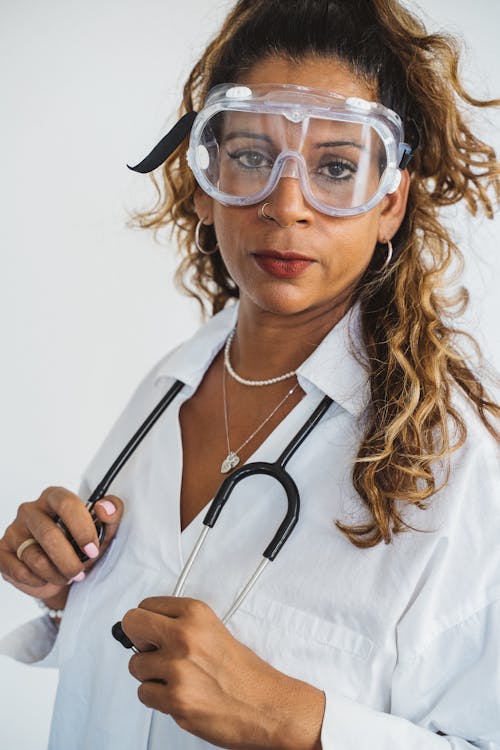 Woman Standing in White Lab  Coat Wearing Safety Glasses and Holding a Stethoscope Draped Around Her Neck