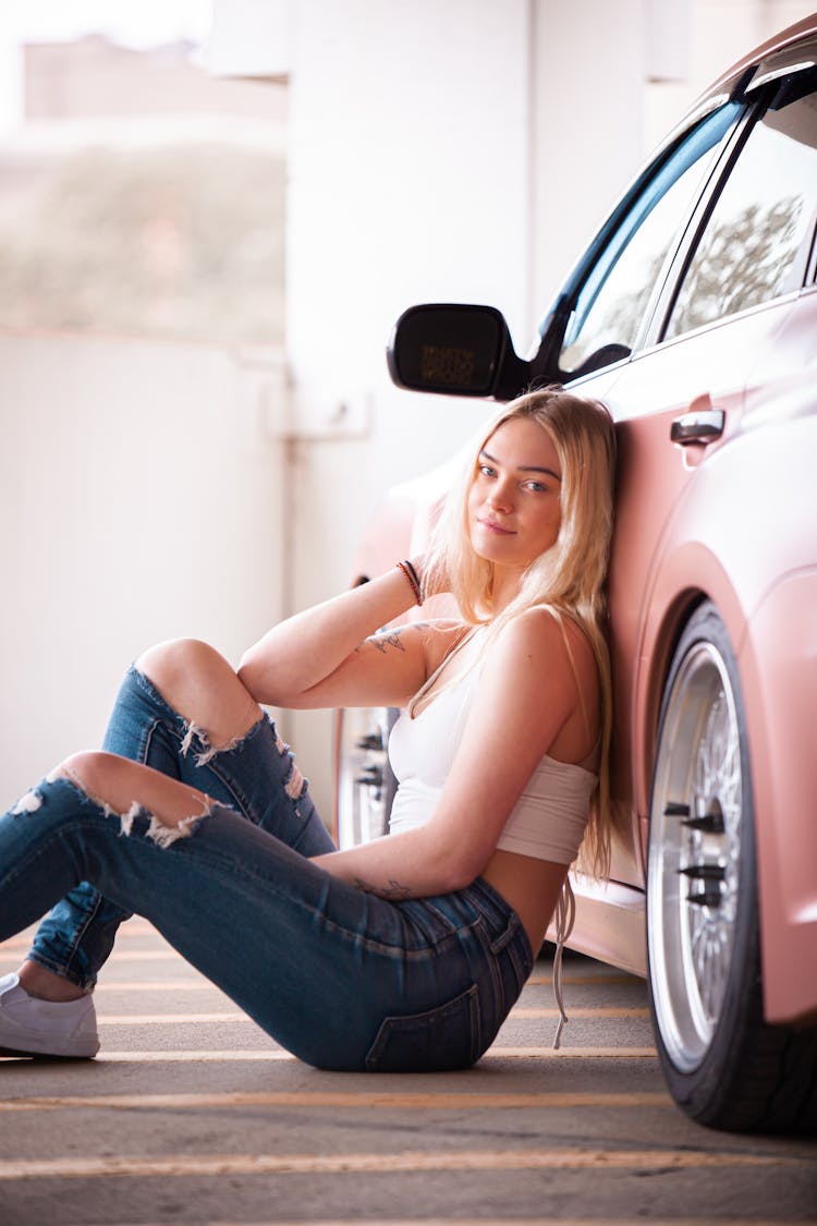 A Woman In Denim Jeans Sitting On The Floor While Leaning On The Car