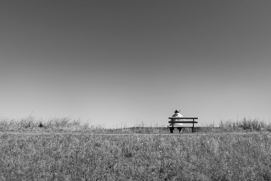 Black and white photo of a lone person sitting on a bench in a grassy field under an open sky.