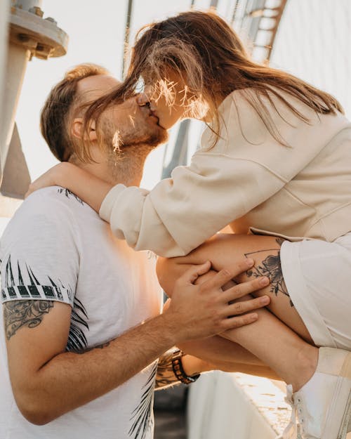 Side view of romantic couple with tattoos in casual clothes standing on street and kissing against sunlight in daytime