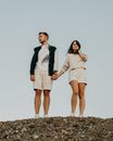 From below full body of confident couple in trendy outfits standing on ground while holding hands under cloudless sky