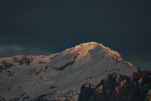 Picturesque view of high mountain slope covered with snow under cloudy dark blue sky in sunset