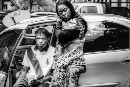 一张黑白照片中，两个人在城市环境里坐在车旁