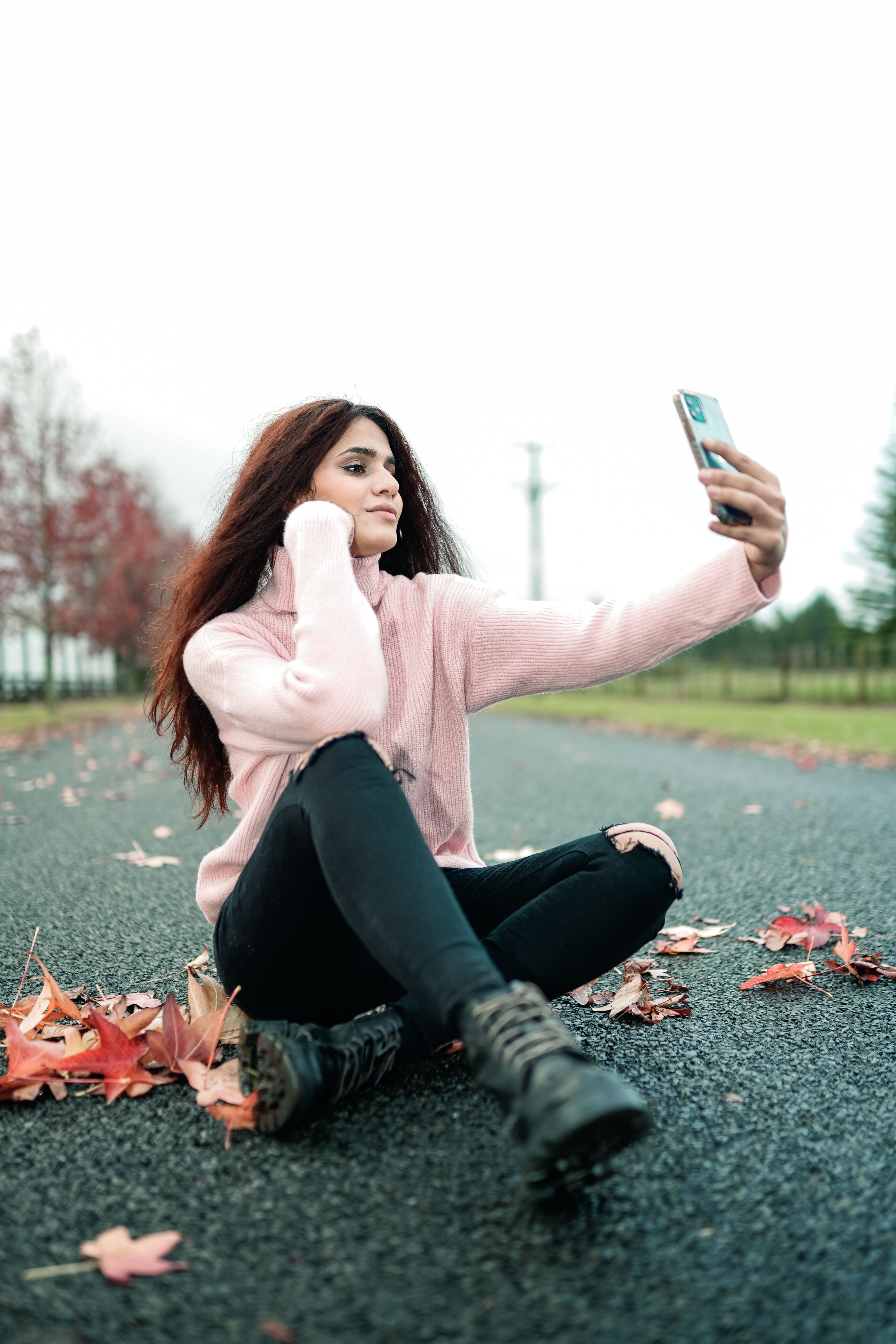 Woman in pink sweater taking a selfie on a leaf-strewn road in autumn.
