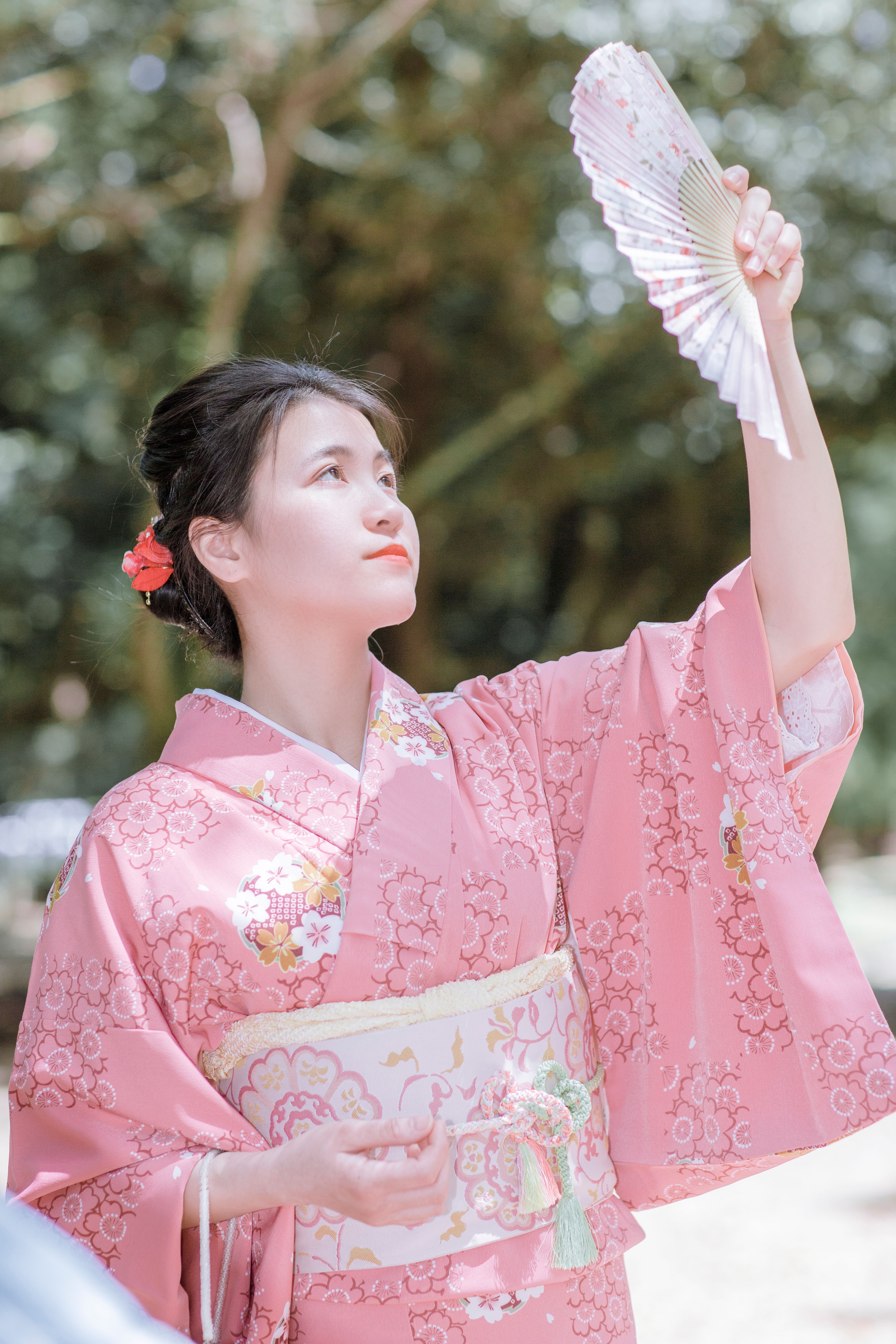 An elegant woman in a pink kimono holding a fan outdoors on a sunny day.