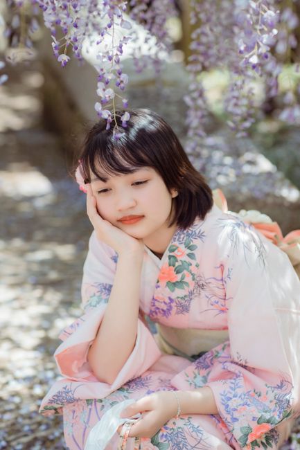 Asian woman in pink kimono surrounded by blooming wisteria flowers, in a peaceful garden setting.