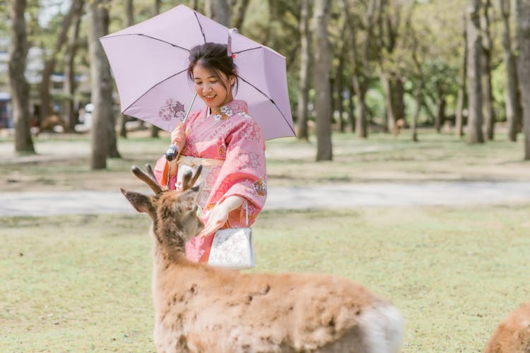 A Woman In Pink Robe Holding An Umbrella While Petting A Deer