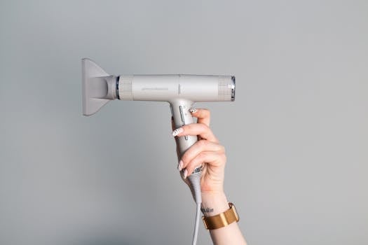 A close-up of a hand with manicured nails holding a sleek hair dryer against a neutral background.